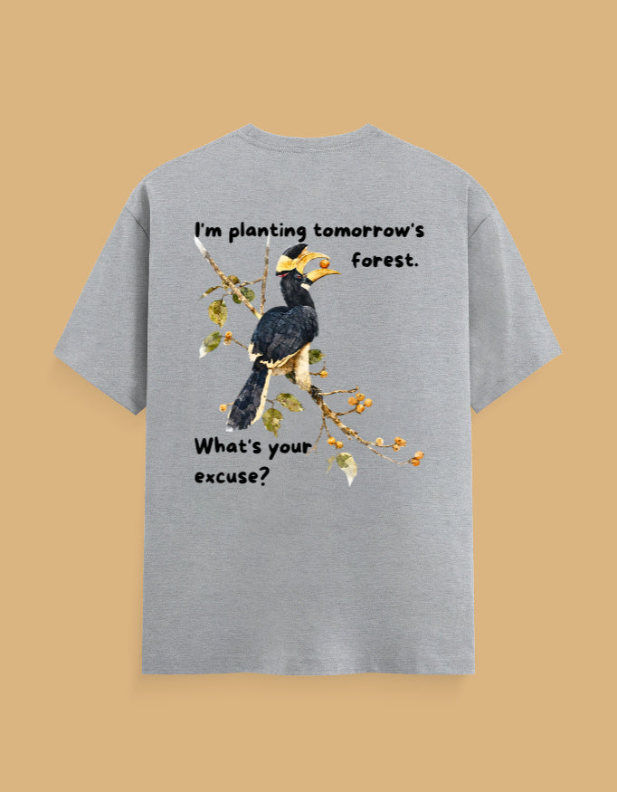 A light heather gray t-shirt shown from the back, featuring a graphic of a hornbill bird on a branch with the text 'I'm planting tomorrow's forest. What's your excuse?' in black lettering, set against a solid tan background.