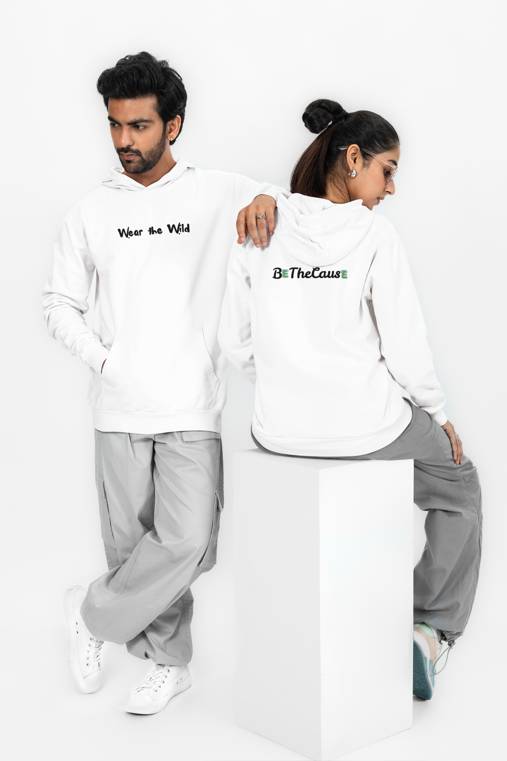 A man and a woman modeling matching white hoodies in a studio setting. The man stands on the left wearing a white hoodie with black text reading 'Wear the Wild' and light gray sweatpants.